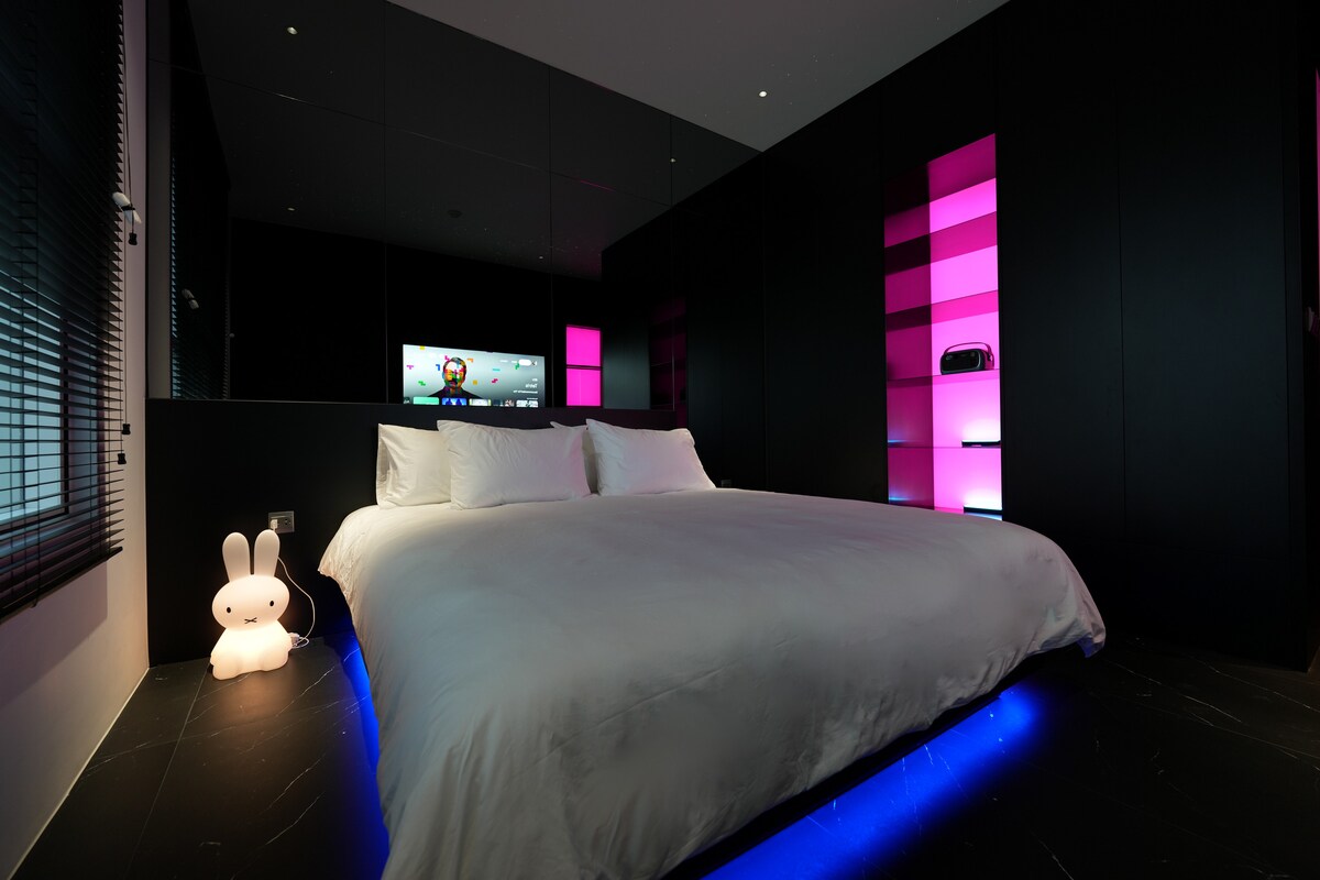 A modern bedroom features a spacious bed with crisp white linens, accentuated by illuminated shelves in pink hues. A flat-screen TV is mounted on the wall, reflecting the elegant black cabinetry. A softly glowing figure adds a touch of warmth to the contemporary design.