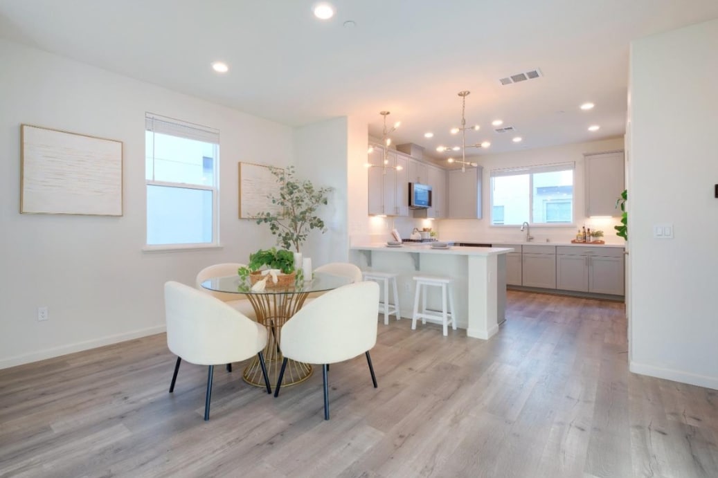 A modern kitchen and dining area features a round table with two cushioned chairs. Natural light enters through two windows, illuminating the space. A kitchen island is visible, complemented by pendant lighting. Light-colored cabinetry and appliances create a clean and inviting environment.