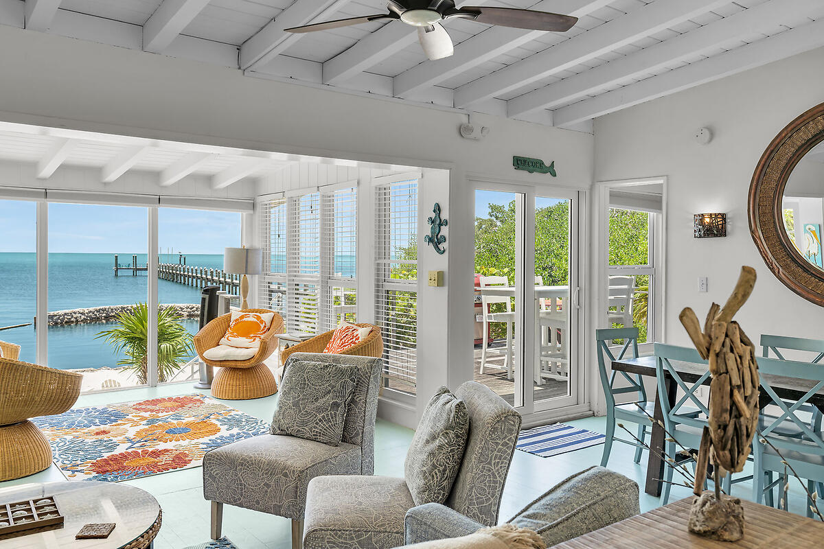 The airy living space features large windows that offer panoramic views of the Gulf. A cozy seating area with woven chairs and a colorful rug is complemented by a dining table set for six. Tropical decor accents the room, enhancing the inviting ambiance.
