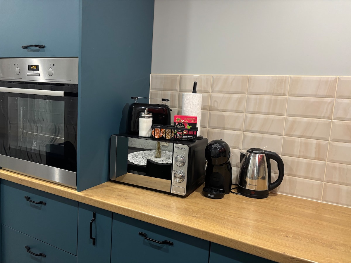 A modern kitchen workspace features sleek cabinetry in deep blue. An array of small appliances is arranged on the wooden countertop, including a microwave, coffee maker, and kettle. Light-colored tiles create a warm backdrop, complemented by a roll of paper towels.