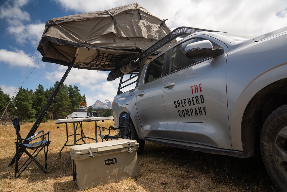 A 4x4 vehicle with a rooftop tent is parked in an open field. A spacious awning extends from the tent, providing shade. A folding table and two chairs are set up nearby, along with a cooler, all surrounded by scenic greenery and mountains in the background.