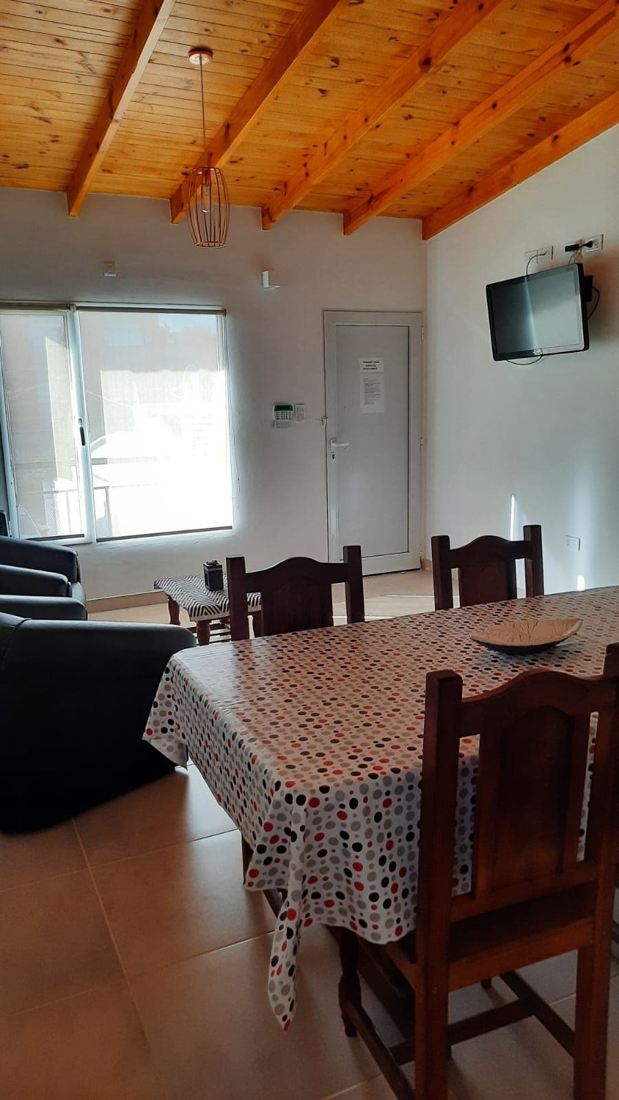A dining area features a table with a colorful tablecloth, surrounded by wooden chairs. The room is brightened by natural light from large glass doors. A small television hangs on the wall, and a door leads to an outside area.