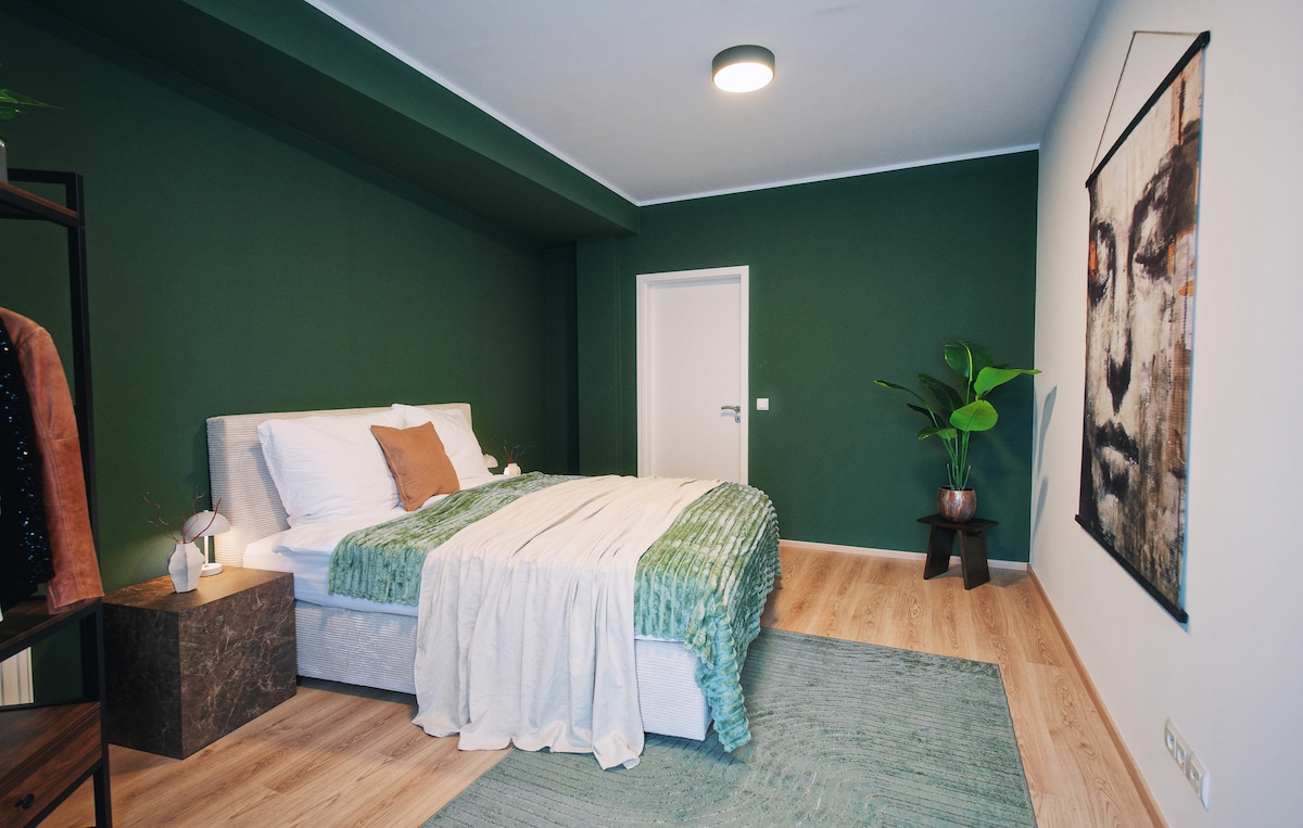 A serene bedroom features a spacious bed adorned with a textured blanket and a decorative pillow, complemented by a green accent wall. A large art piece hangs above the bed, and a leafy plant adds a natural touch to the room. Light hardwood flooring enhances the ambiance.