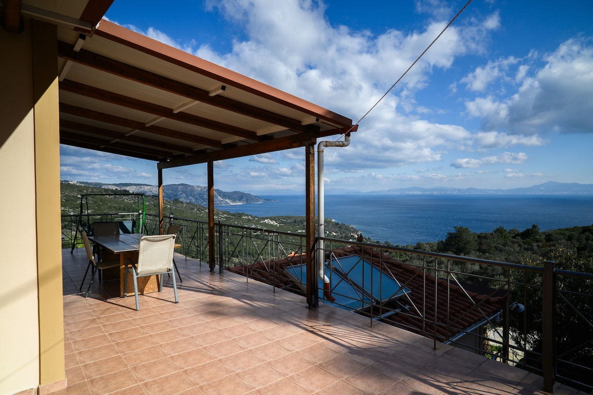 A spacious outdoor terrace features a sturdy wooden table with accompanying chairs, offering panoramic views of the sea and surrounding landscape. The area is partially shaded by a roof, with sunlight illuminating the tiled floor, creating an inviting space for relaxation.