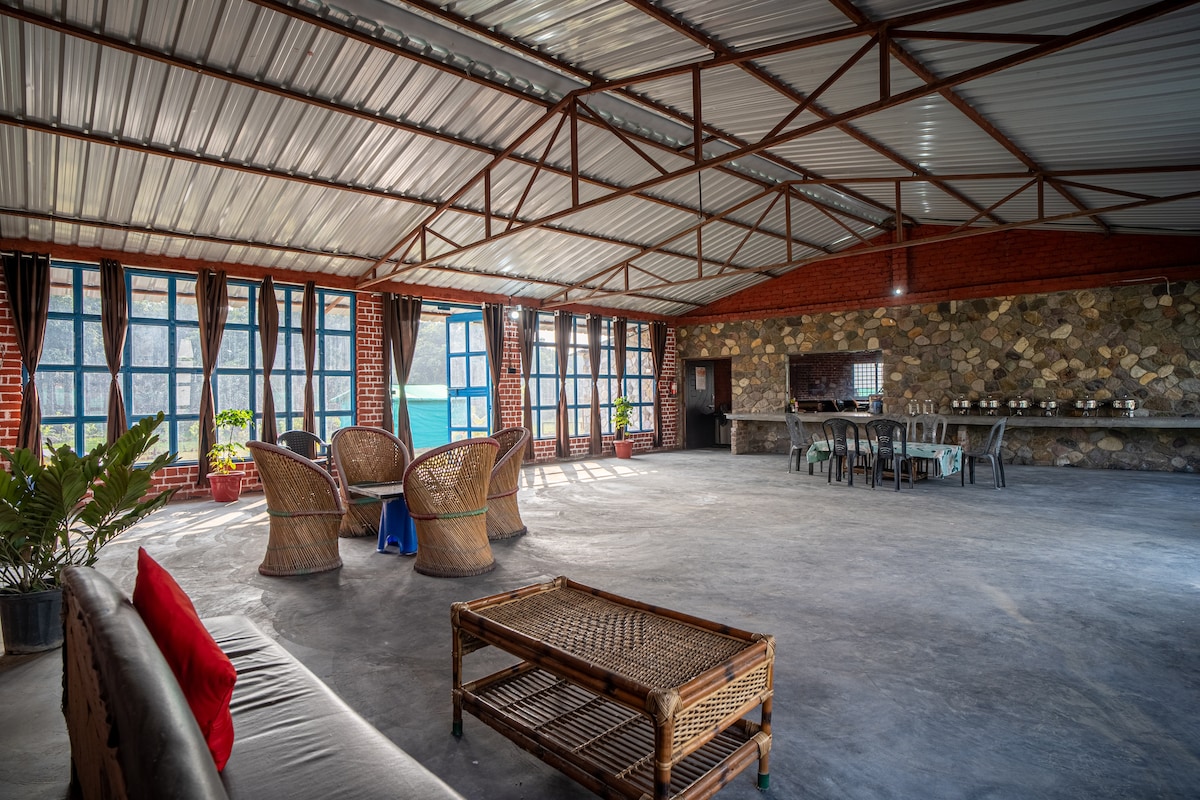 A spacious common area is characterized by large windows allowing natural light to fill the space. Several wicker chairs and a coffee table are arranged for relaxation, while stone walls provide a rustic backdrop. A minimalistic bar area is visible in the distance.