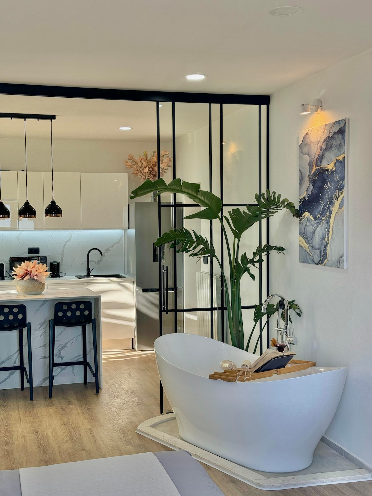 An elegant bath area features a freestanding white bathtub complemented by a wooden tray holding wine and glasses. Behind it, a fully equipped kitchen is visible, with a marbled backsplash and modern bar stools. Large windows allow natural light to illuminate the space.