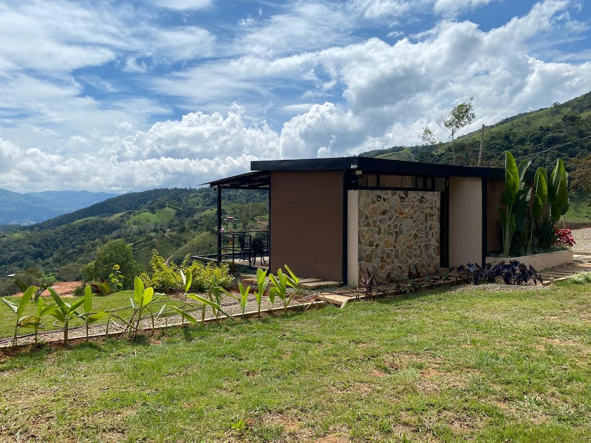 A modern cabin is set against a backdrop of rolling hills. The structure features a combination of stone and smooth walls, with large windows allowing for natural light. Surrounding greenery includes well-maintained grass and various plants, enhancing the tranquil outdoor setting.