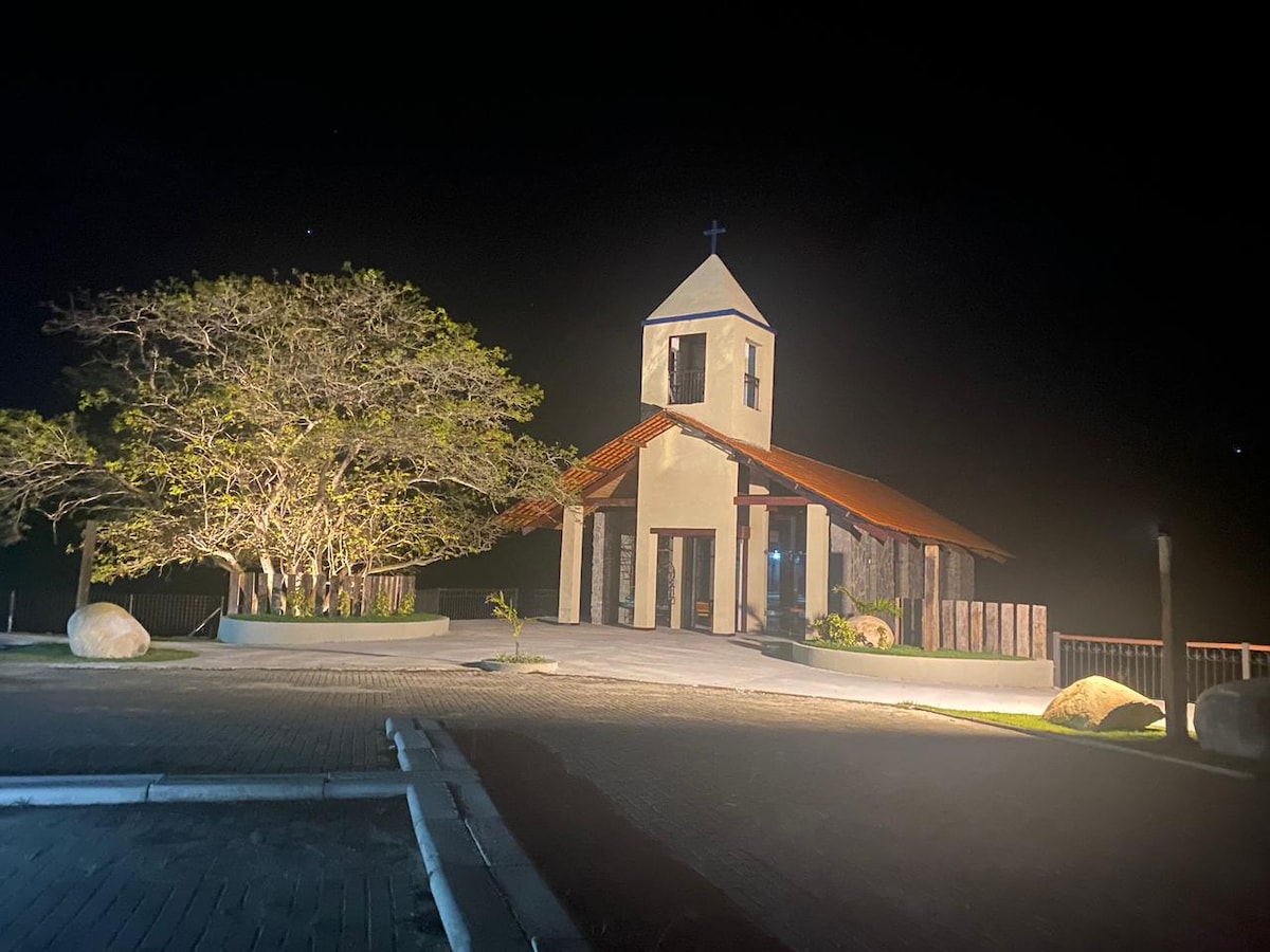 A charming chapel is shown at night, with soft lighting illuminating its façade. A large tree stands nearby, complementing the serene environment. The building features a pointed roof adorned with a cross, and the pathway is lined with large stones, creating a welcoming atmosphere.
