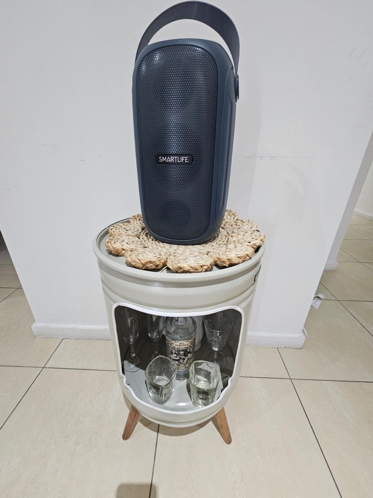 An elegant storage unit with wooden legs showcases a collection of glassware inside. A textured navy beverage cooler rests on top, accompanied by a woven decorative mat. The overall presentation emphasizes functional and stylish organization within the living space.