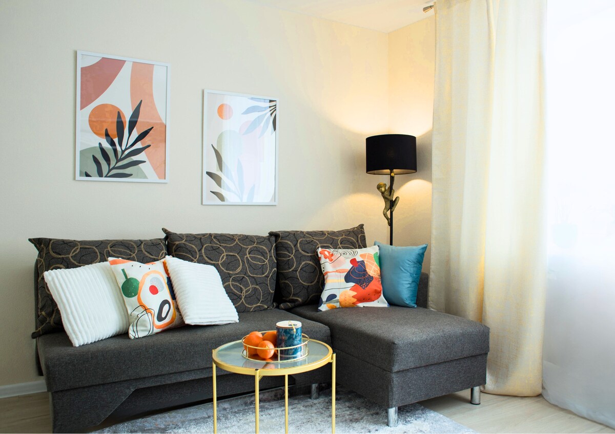 A contemporary living area features a dark gray sectional sofa adorned with a variety of decorative cushions. A round, golden coffee table sits nearby, displaying fresh fruit. Bright artwork is displayed on the walls, and natural light filters in through a sheer curtain.