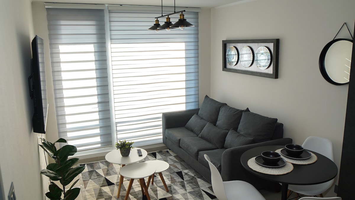 A cozy living area features a gray sofa positioned in front of a wall-mounted television. A round white table is set with dark dishes, accompanied by four sleek chairs. Natural light filters through layered window shades, illuminating the geometric-patterned rug and a small potted plant.