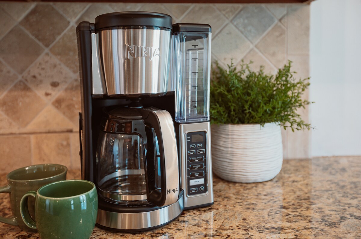 A coffee maker is positioned on a granite countertop, with a glass carafe and control panel clearly visible. Two green mugs rest beside it, and a textured white planter with greenery adds a touch of color to the scene.