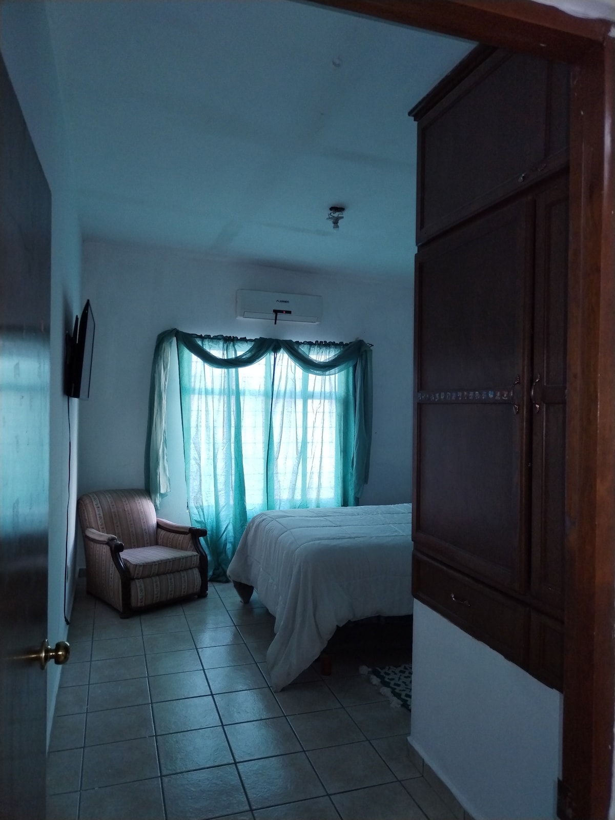 A serene bedroom is visible with a spacious bed dressed in white linens. An armchair sits in one corner, complemented by sheer turquoise curtains that softly diffuse natural light. A small air conditioning unit is discreetly mounted on the wall, enhancing comfort.