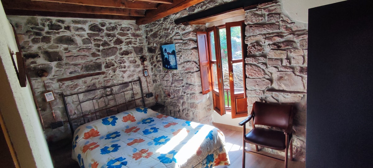 A charming bedroom features exposed stone walls and wooden beams overhead. A spacious bed with a patterned quilt is set against the backdrop of a natural light-filled window, which opens to the outside. A leather chair is positioned in the corner for relaxation or reading.