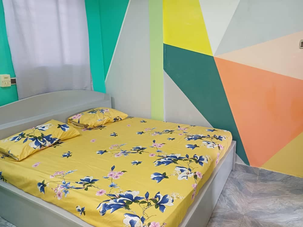 A spacious sleeping area is highlighted by a large bed with a bright yellow floral bedspread and matching pillows. The walls feature a mix of vibrant colors, creating an energetic yet calming atmosphere. Natural light enters through a nearby window, enhancing the space.