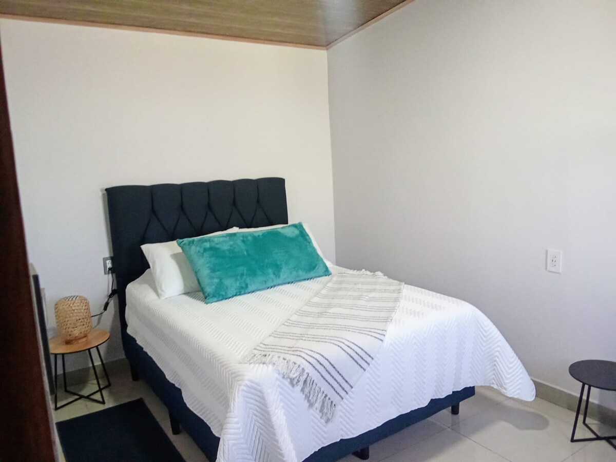 A cozy sleeping area features a spacious bed with a tufted headboard, topped with a turquoise accent pillow. A lightweight blanket drapes over the bed, while a small side table and a minimalist stool are positioned beside it.