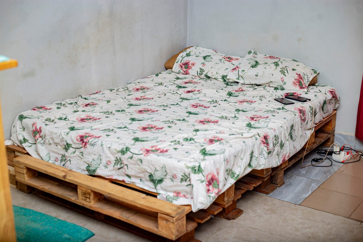 A spacious bed with a floral-patterned comforter is positioned on a wooden pallet base. Soft pillows are arranged at the head of the bed, while a small electronic device is placed on the side. The surrounding walls feature a light-colored finish.