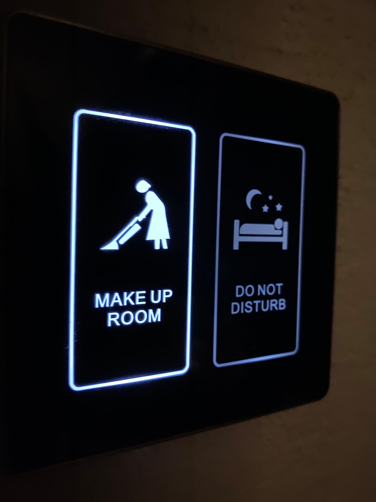 A wall-mounted sign is displayed, featuring illuminated symbols for room service options. The left side indicates 'Make Up Room' with a figure vacuuming, while the right side shows 'Do Not Disturb' with an image of a bed and stars.