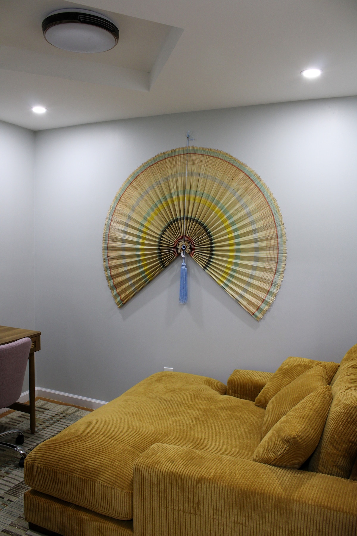 A cozy seating area features a soft, mustard-yellow couch positioned against a light grey wall. Above the couch, a large decorative fan with a mix of beige, green, and blue tones adds a vibrant focal point to the room. Soft overhead lighting illuminates the space.