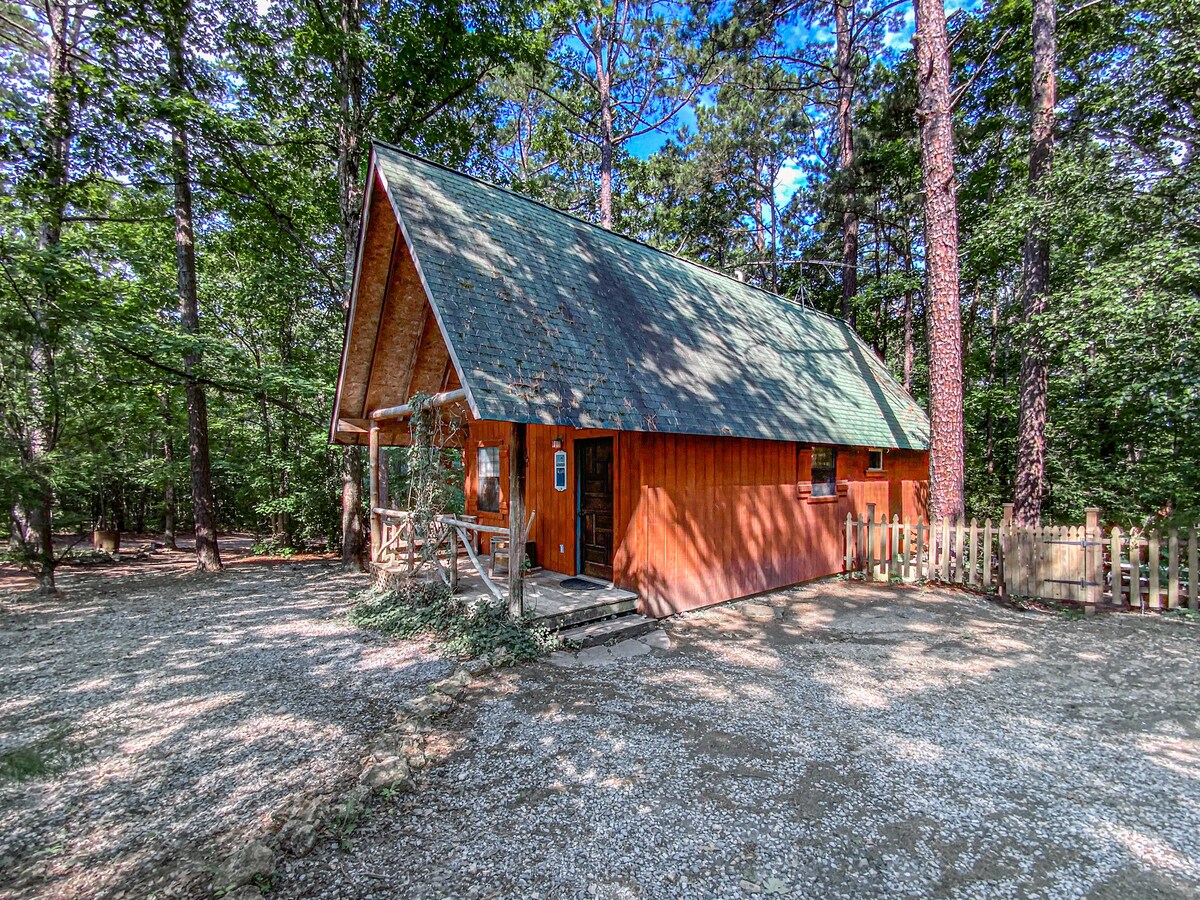 A quaint cabin with a sloped roof stands nestled among tall trees. The exterior is composed of warm wood tones, complemented by a green shingled roof. A gravel path leads to the entrance, lined by a rustic fence that enhances the natural setting.