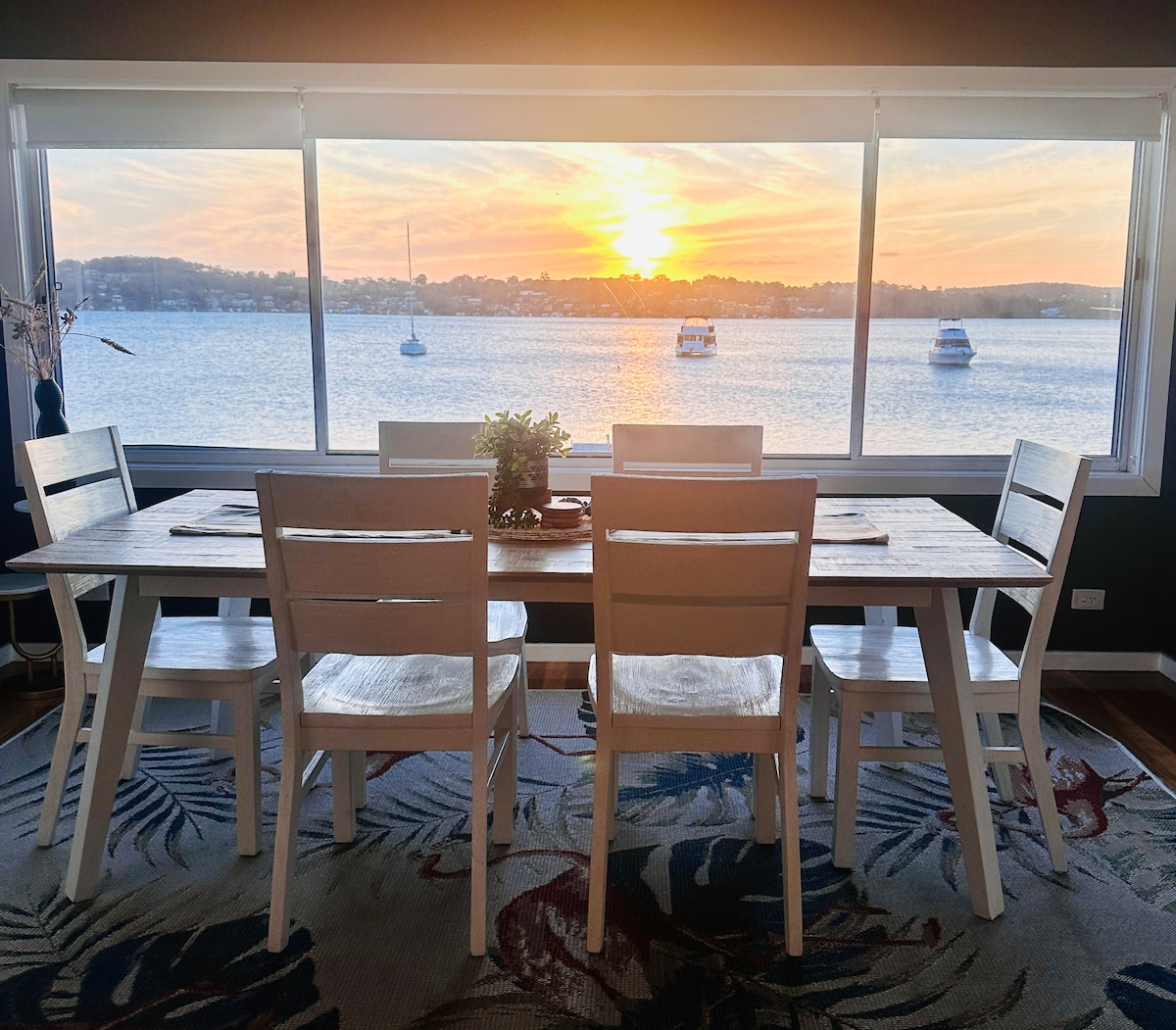 A dining area is shown with a large window framing a sunset view over the water. A wooden table is surrounded by six white chairs, and a small plant sits at the center. The rug features a colorful pattern, adding warmth to the space.