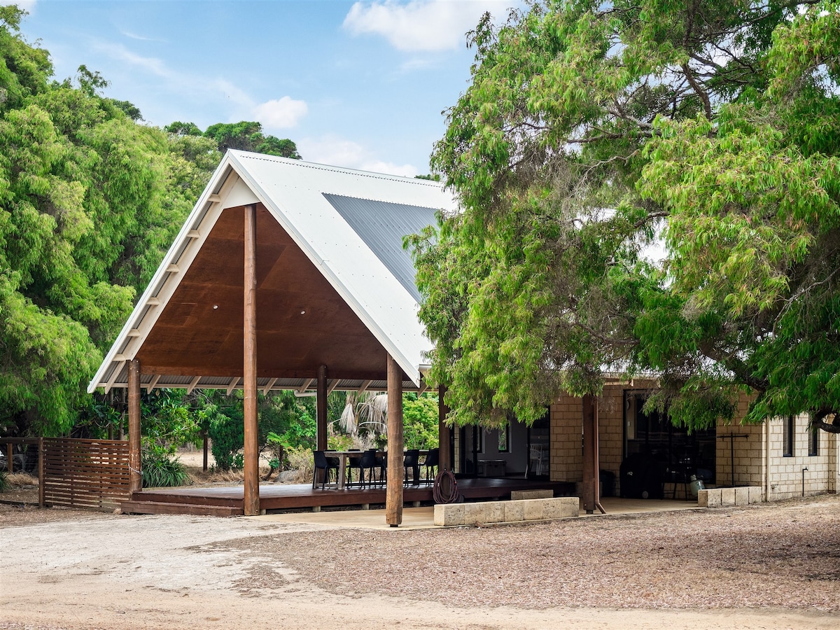 The spacious front deck of the Farm Guest House is sheltered by a sloped roof supported by wooden posts. Outdoor seating is arranged around a large dining table, surrounded by trees and greenery, blending indoors and outdoors in a tranquil environment.