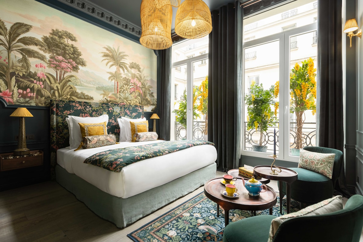 A spacious bedroom features a king-size bed adorned with patterned linens and decorative pillows. Large windows provide views of greenery outside, while textured curtains frame the scene. Two small tables hold colorful tea sets, contributing to the room's elegant decor.