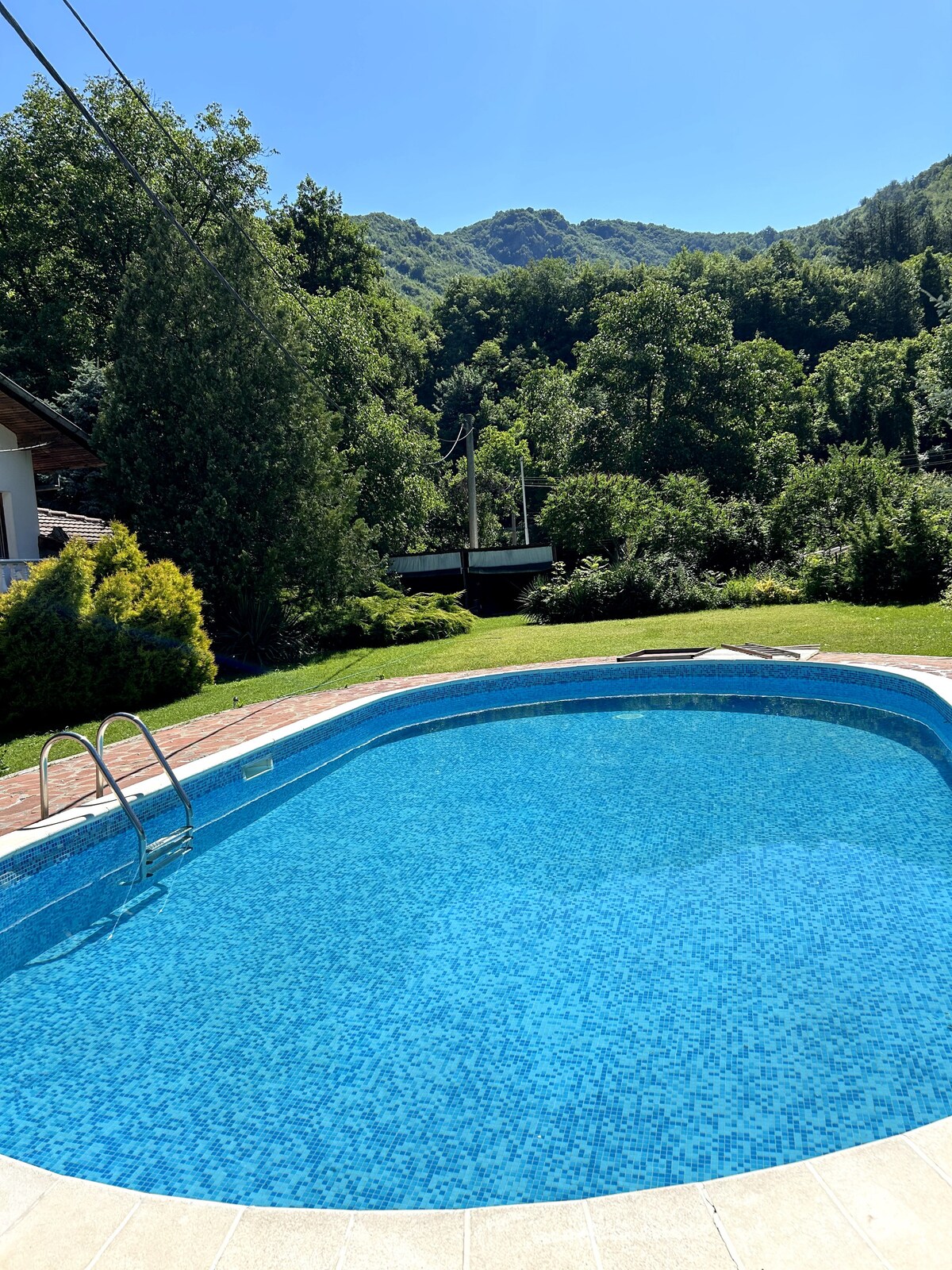 A clear, inviting swimming pool reflects the bright blue sky, embraced by lush green trees and surrounding hills. The pool area features a gentle slope leading to the water and is framed by well-cared-for landscaping offering a tranquil view of nature.