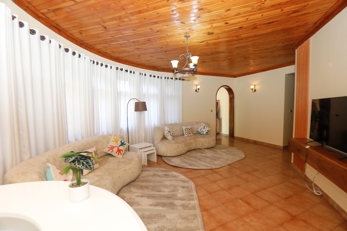 A circular living area features two plush, curved sofas adorned with decorative cushions. Soft white curtains frame large windows, allowing natural light to fill the space. A wooden ceiling enhances the cozy feel, while a sleek television sits on a wooden console opposite the seating.