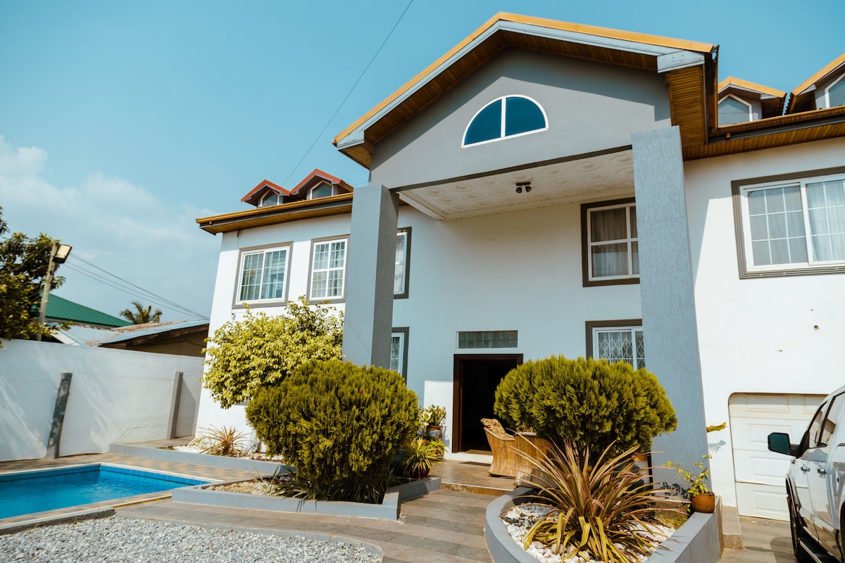A modern two-story home is depicted, featuring a prominent entrance framed by a large arched window. Lush green bushes and well-maintained landscaping surround the space, with a private swimming pool visible to one side, reflecting the clear blue sky.