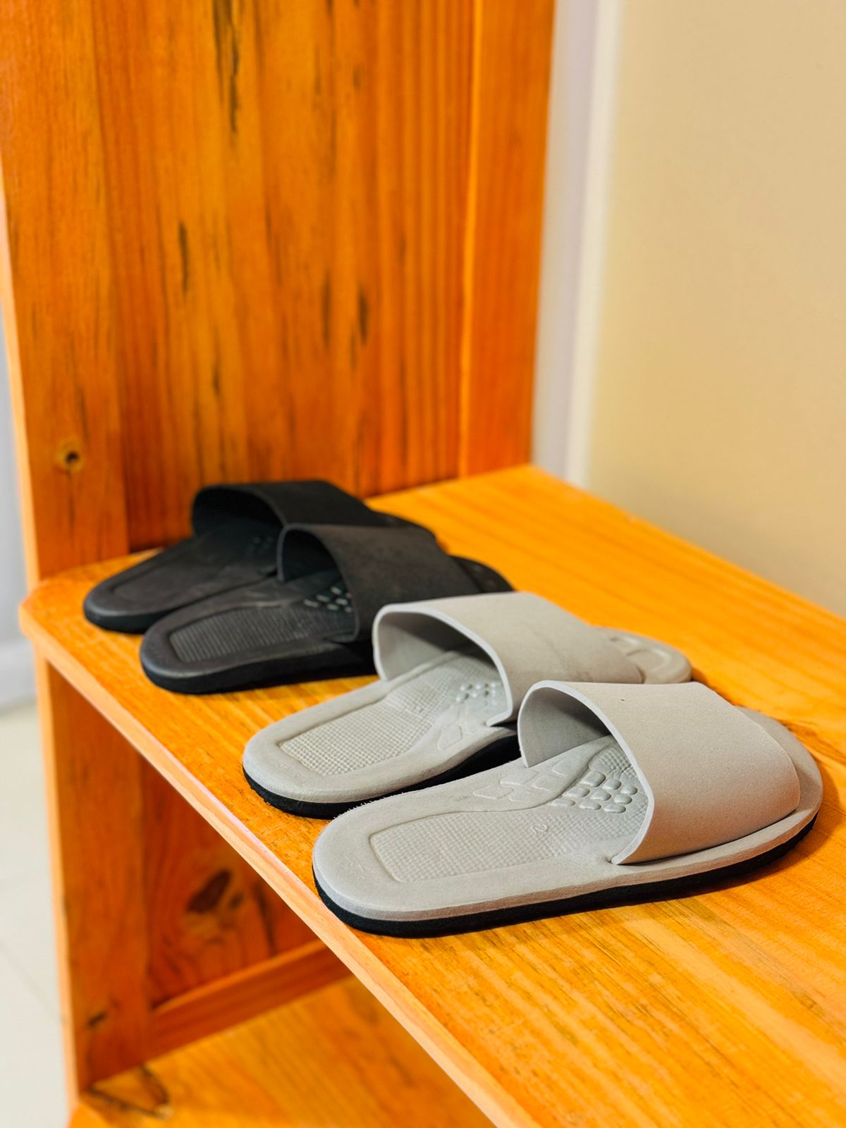 A pair of black and gray slippers are arranged neatly on a wooden shelf. The slippers are designed for comfort, featuring soft straps and textured soles. The warm wood grain of the shelf provides a natural backdrop.