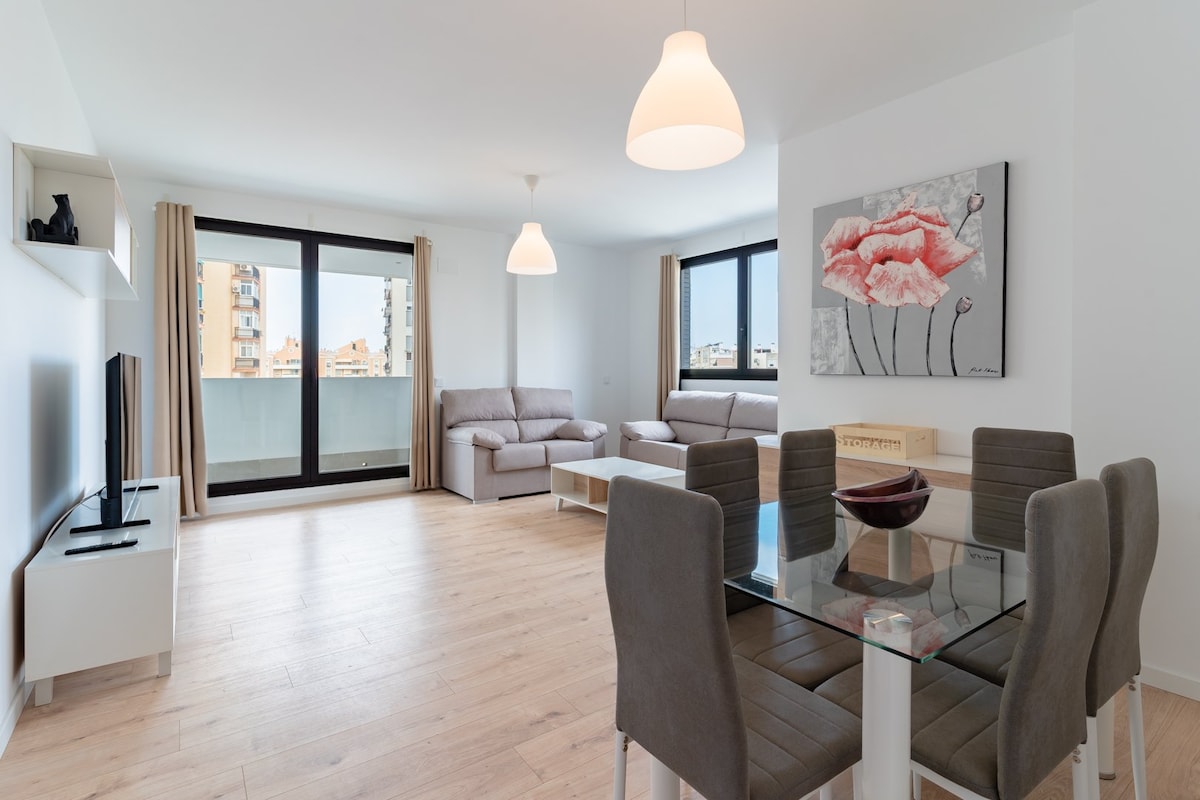 A bright and airy living area is presented with large windows allowing natural light to fill the space. A comfortable sofa is positioned alongside a glass dining table surrounded by grey chairs. A piece of artwork featuring red flowers adds a touch of color to the minimalist decor.