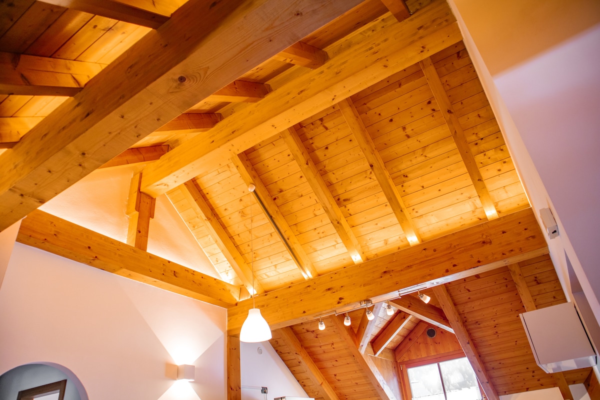 The image captures the wooden ceiling of the loft, featuring exposed beams that create a spacious atmosphere. Soft lighting along the beams enhances the natural wood tones, while large windows allow natural light to illuminate the area, contributing to a warm environment.