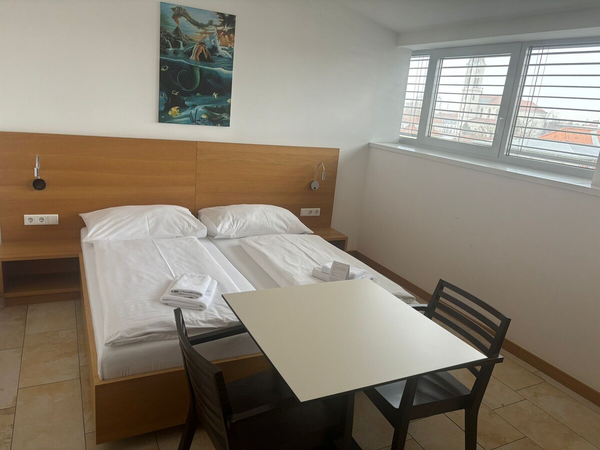 A bright sleeping area features a spacious bed with neatly arranged white linens. A small table with two chairs occupies the corner, while a colorful artwork adorns the wall above the bed. Natural light filters through horizontally placed windows.
