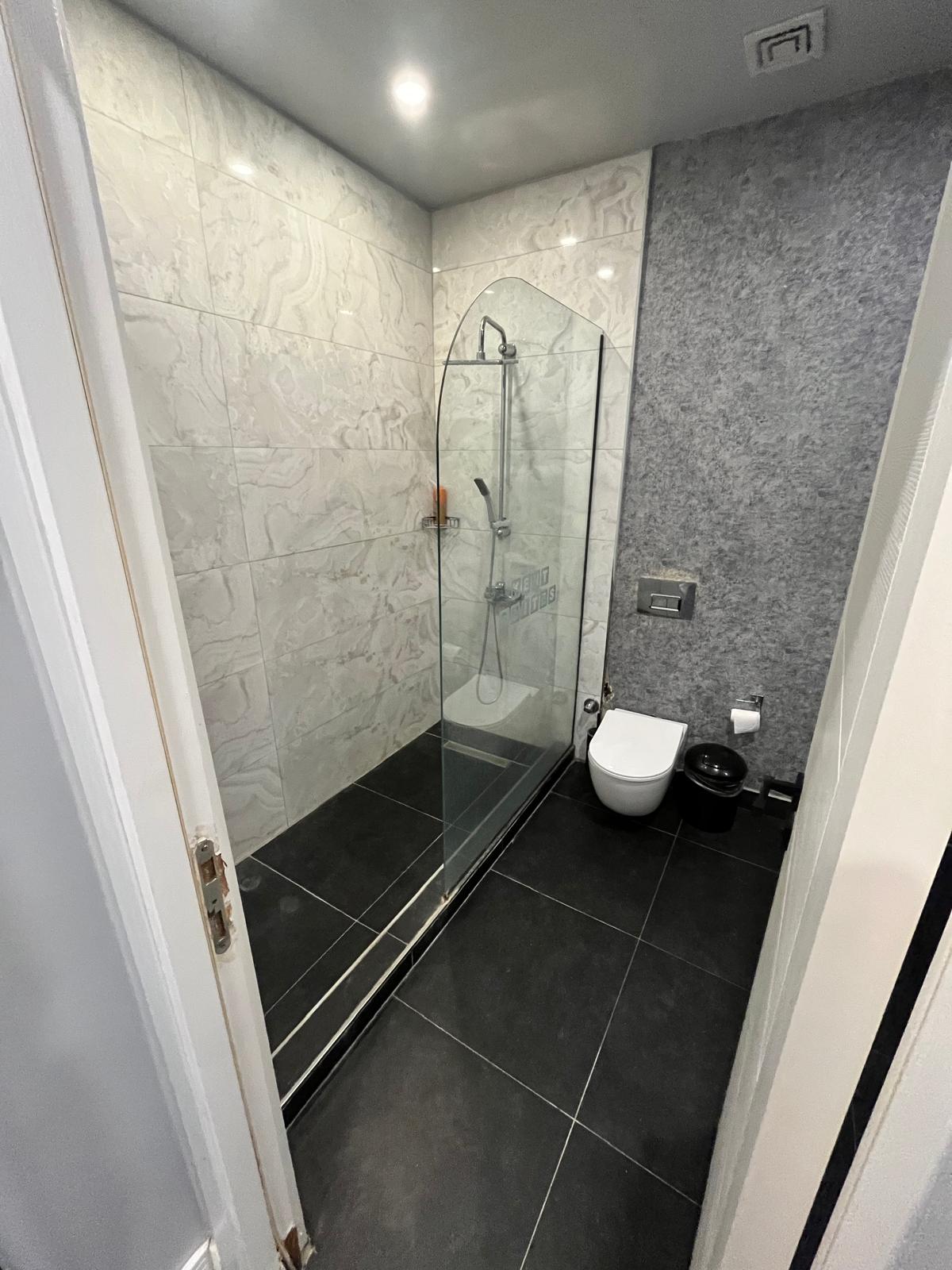 A modern bathroom is equipped with a glass shower enclosure featuring a rainfall showerhead. The sleek black floor contrasts with the light-colored tiled walls. A wall-mounted toilet is positioned beside the shower, and a brushed metal faucet complements the overall design.