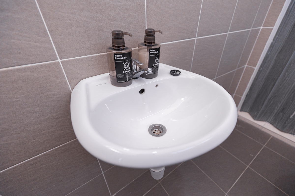 A clean, modern sink is positioned against a tiled wall. Two labeled soap dispensers are placed on the sink's edge. The floor is covered in dark tiles, providing a contemporary contrast to the light-colored wall tiles.