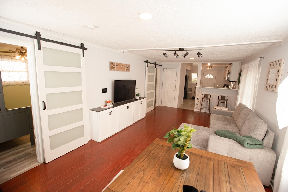 A well-lit living area features a spacious gray sofa, a wooden dining table, and dual sliding barn doors. A television is mounted on the wall, accompanied by a modern white sideboard. Natural light enters through large windows, enhancing the warm ambiance of the space.