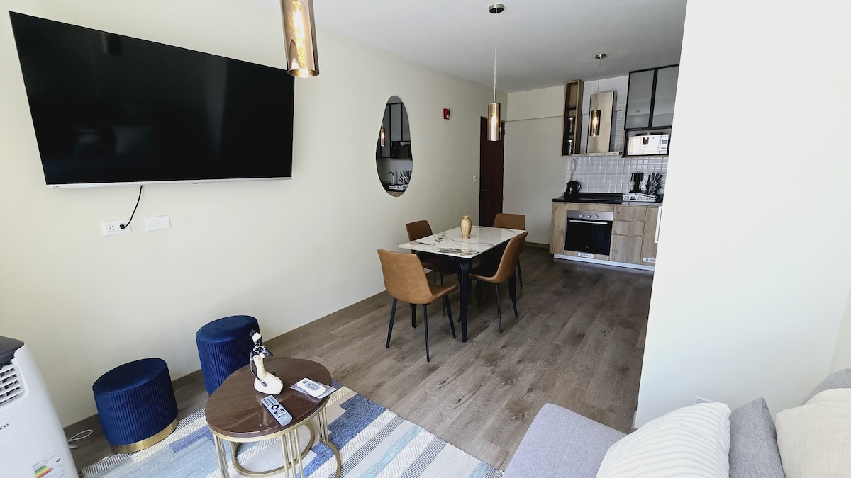 A modern living area is presented, featuring a wall-mounted television and an elegant dining table with four chairs. A stylish mirror and a cozy sofa are also visible, along with decorative elements on a coffee table. Soft lighting is provided by pendant fixtures.