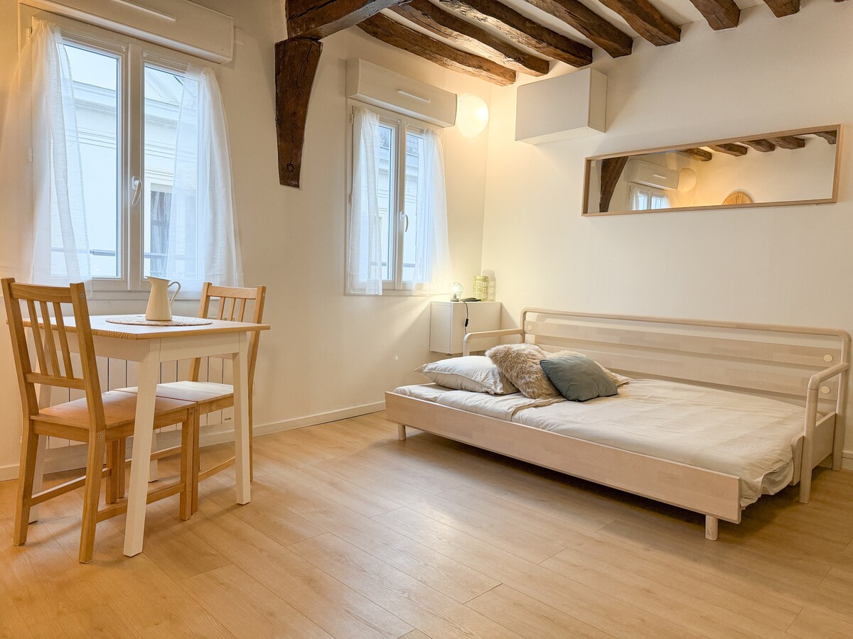 The studio features warm wooden beams and light-toned flooring. A compact table with two chairs is positioned next to a large window, which allows natural light to fill the space. A foldable bed rests against the wall, adorned with soft cushions.