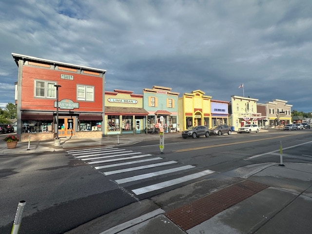 A row of colorful storefronts lines the street, showcasing varied architectural styles. The buildings feature large windows and inviting entryways, with a wide crosswalk visible in the foreground. Cloudy skies create a serene atmosphere, contributing to the charm of the town.