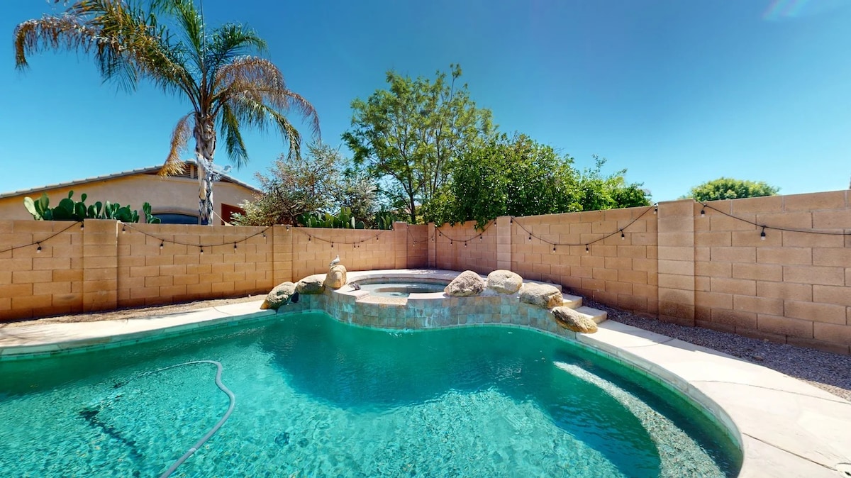 A serene outdoor space features a private pool with gentle curves, surrounded by a natural stone edge. A hot tub is visible beside the pool, with a landscaped yard containing palm trees and lush greenery. Neutral-colored walls create a private and inviting atmosphere.