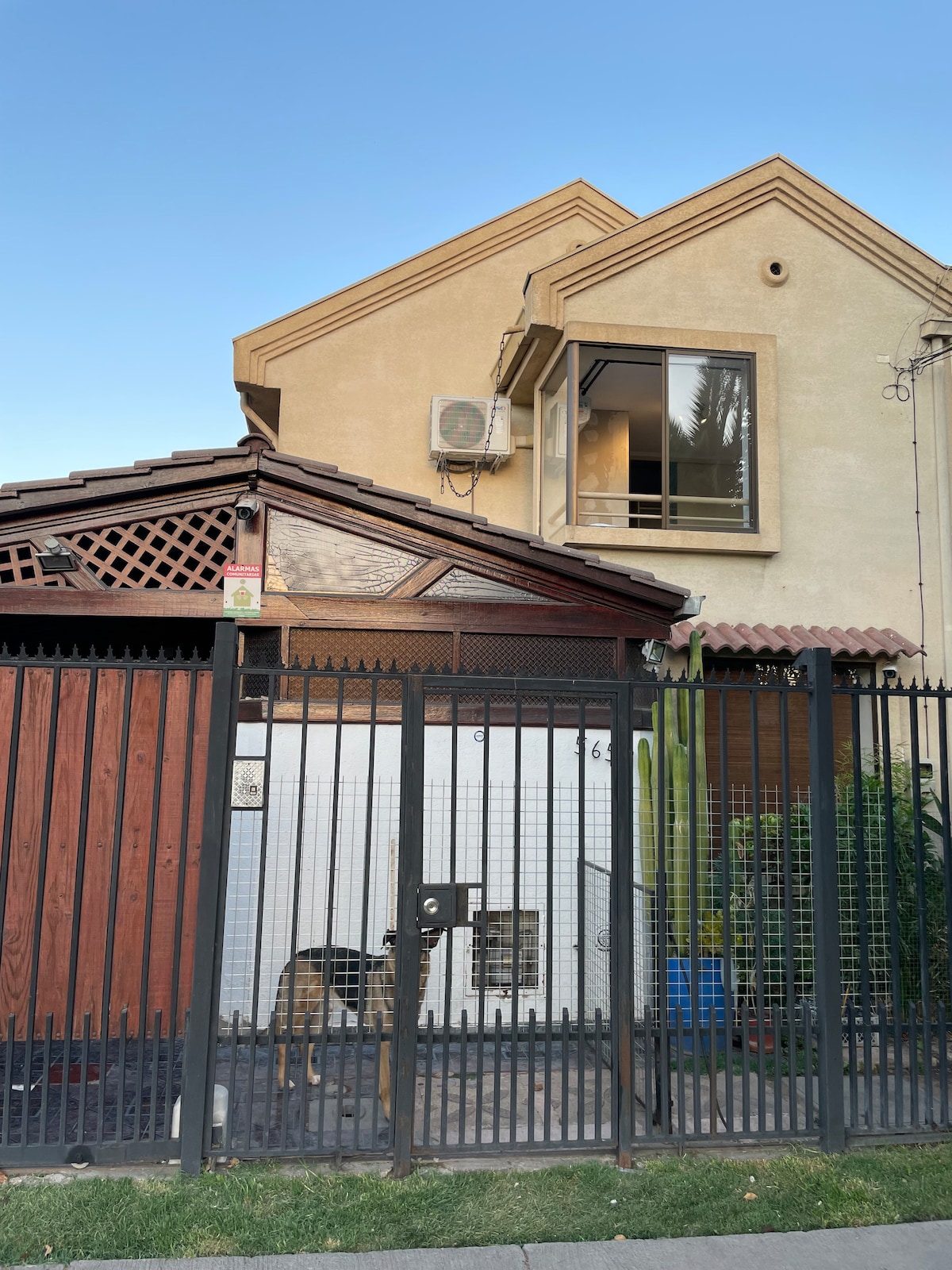 A two-story building features a combination of textured stucco and wood accents. An air conditioning unit is mounted on the exterior wall, while a gated area presents a garden space. The setting is framed by a wrought iron fence, adding to its inviting character.