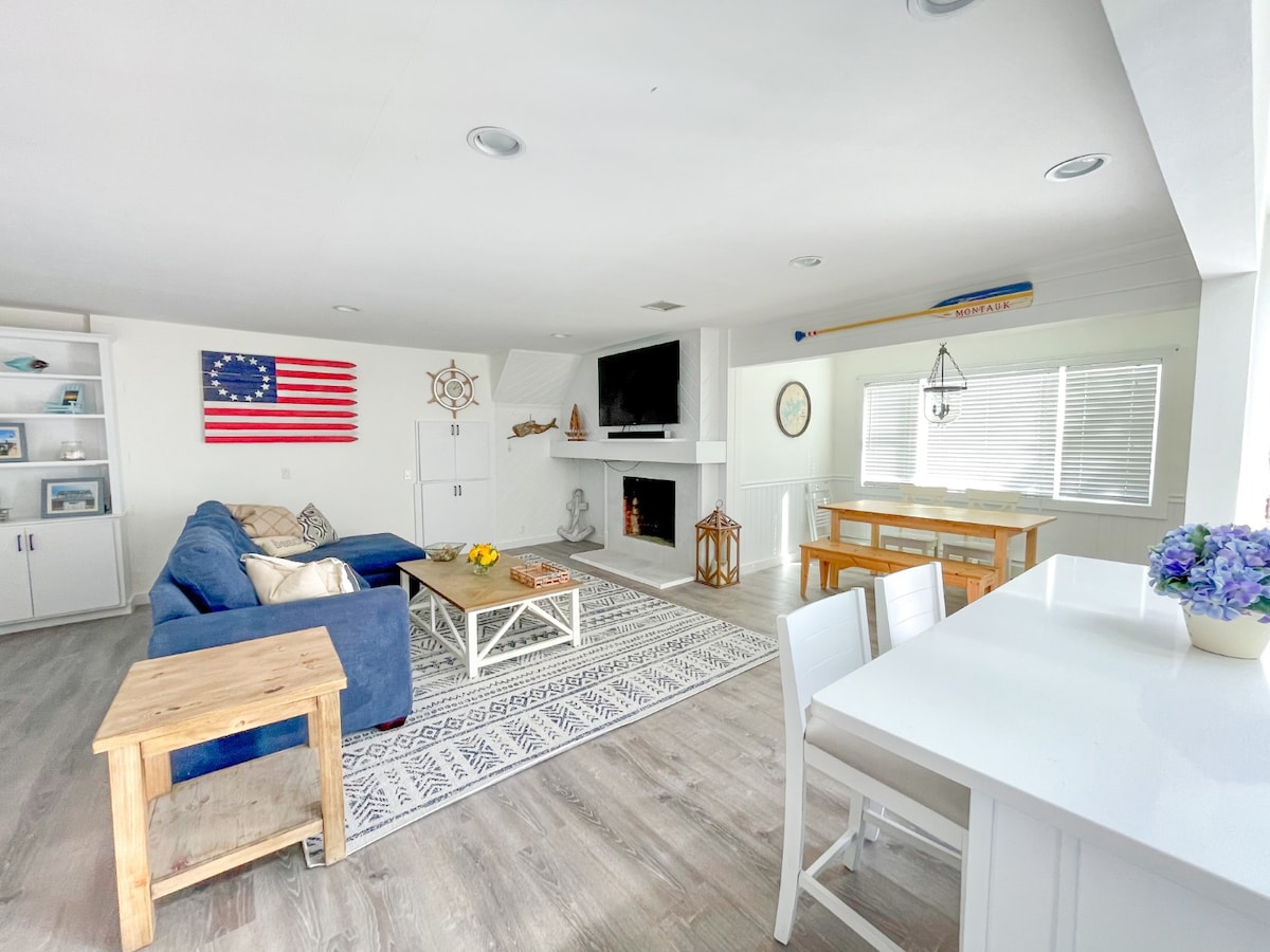 A spacious living area features a comfortable blue sofa accompanied by a wooden coffee table. A decorative area rug covers the floor, while a television is mounted above a white fireplace. A dining table with chairs is positioned near large windows, allowing ample natural light.