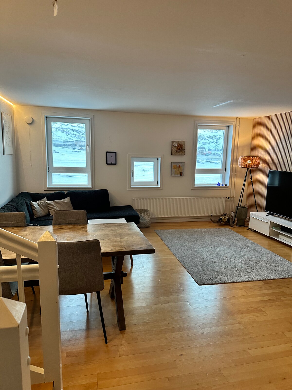 A modern living area is presented with a dark sofa and a small dining table surrounded by four chairs. Natural light enters through three windows, illuminating the wooden floor. A soft rug lies beneath a light fixture and a media unit with a television.