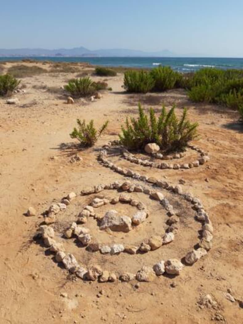 A sandy landscape is highlighted by a spiral formation made of stones, set against a backdrop of the sea and distant mountains. Lush green shrubbery is visible alongside the spiral, contributing to the natural scenery under a clear blue sky.