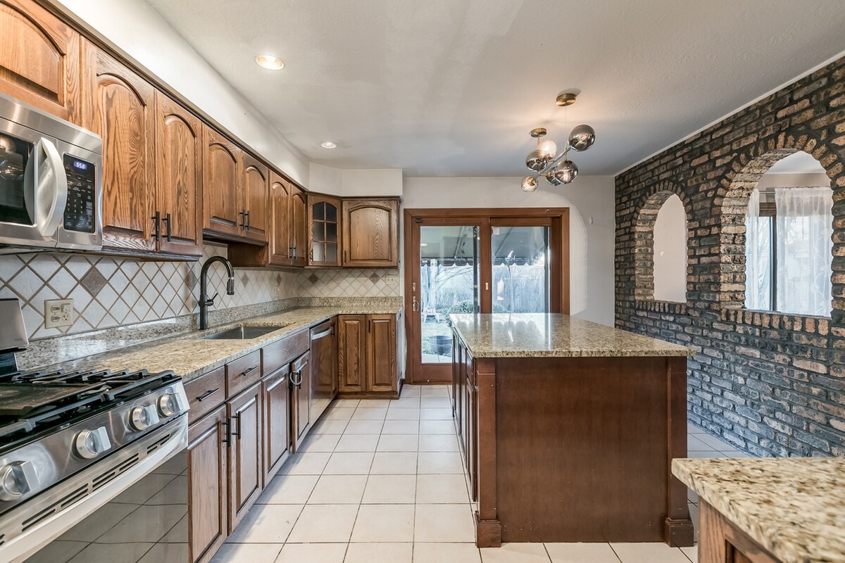 A spacious kitchen is equipped with wooden cabinets and granite countertops. Stainless steel appliances, including a gas stove, are visible. Adjacent to the kitchen is a dining area with a doorway leading to an outdoor space, framed by large glass doors that enhance natural light.