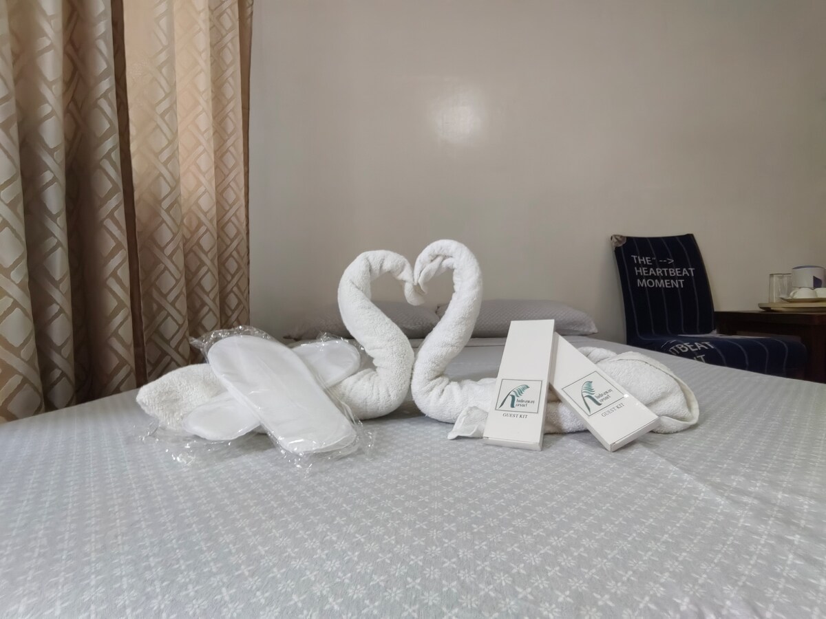 A cozy bed features elegantly twisted white towels shaped like swans, positioned to create a romantic heart formation. Soft slippers and neatly arranged toiletries are placed nearby, complementing the serene decor, while light drapes softly filter through the curtains.