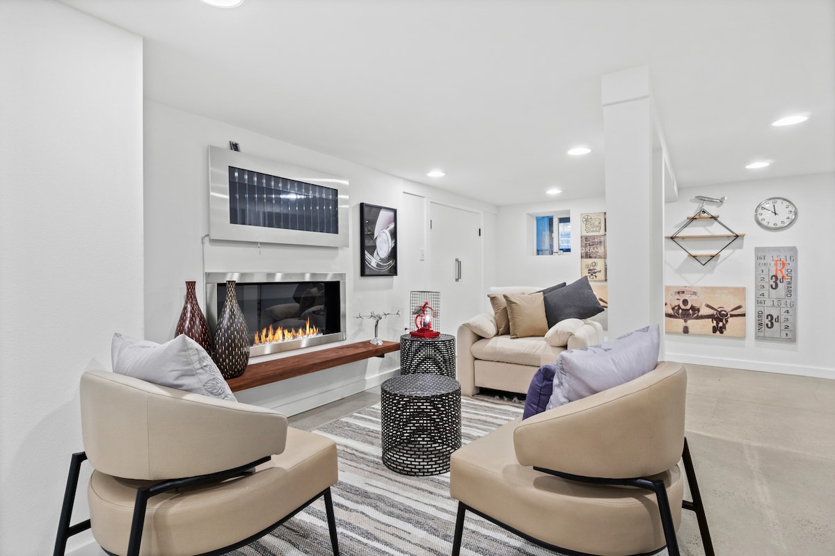 A cozy den area displays two light-colored chairs set around a circular coffee table. A modern fireplace is located on one wall, complemented by framed artwork and a wall clock. Soft lighting enhances the inviting atmosphere, creating a perfect space for relaxation.