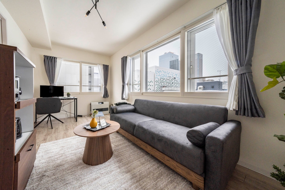 A modern living area featuring a spacious gray sofa and a round wooden coffee table at the center. Large windows allow natural light to fill the room, coupled with elegant curtains. A small workspace with a chair is positioned nearby, enhancing the functionality of the space.