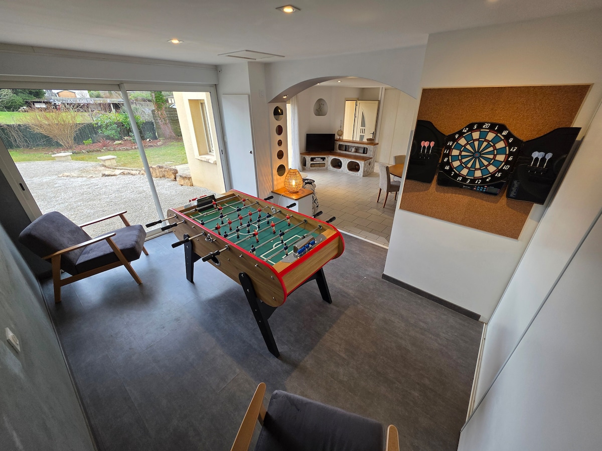 A bright living space features a foosball table as the centerpiece, with a dartboard mounted on a cork surface nearby. Large glass doors open to the outside, offering a view of the garden. A comfortable armchair is positioned to allow relaxed seating.